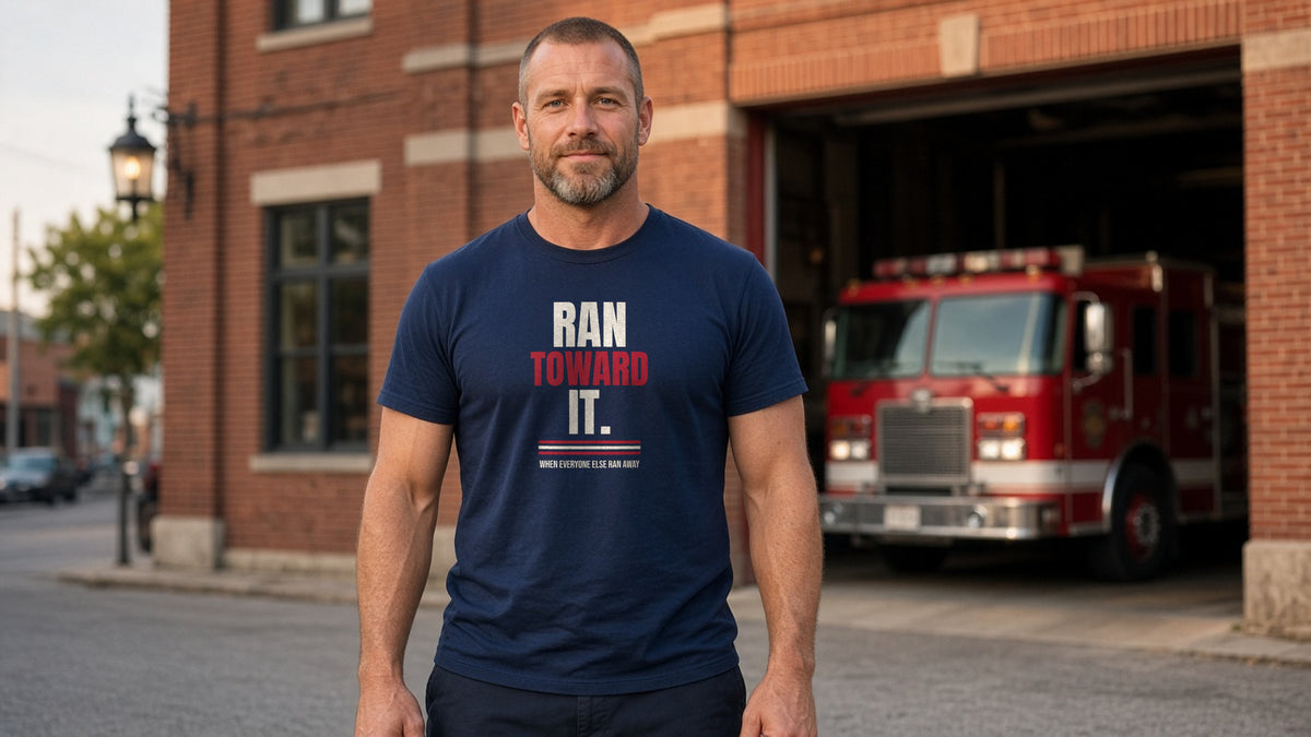 Man in a navy Ran Toward It tee standing outside a brick firehouse with a fire engine in the bay